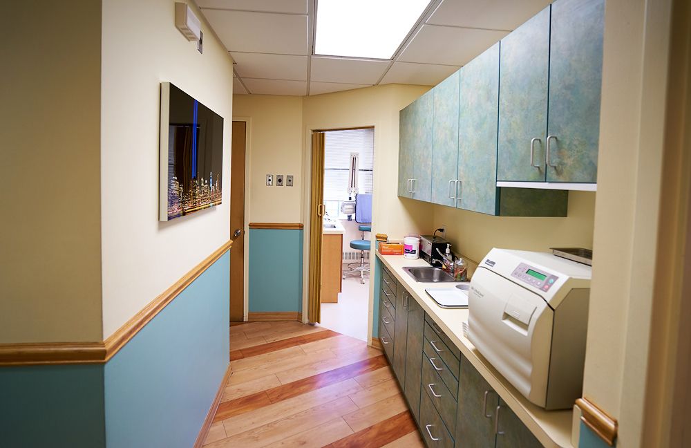 The image depicts an interior space that appears to be a small office or room with walls painted in various shades of blue, green, and cream. There is a desk with a computer monitor on it, along with other items such as a printer and what looks like a cup. A door with a window is visible, and the floor is covered with wood-like tiles. The overall atmosphere suggests a professional environment with a touch of color.