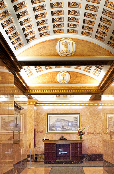 The image depicts a luxurious lobby with a high ceiling, featuring an ornate chandelier, a large vaulted ceiling with intricate designs and gold accents, and decorative moldings. The floor is tiled, and there are framed pictures on the walls. In the foreground, there s a reception desk with a sign that reads  THE DALLAS MORGAN  in capital letters, indicating the name of the establishment.