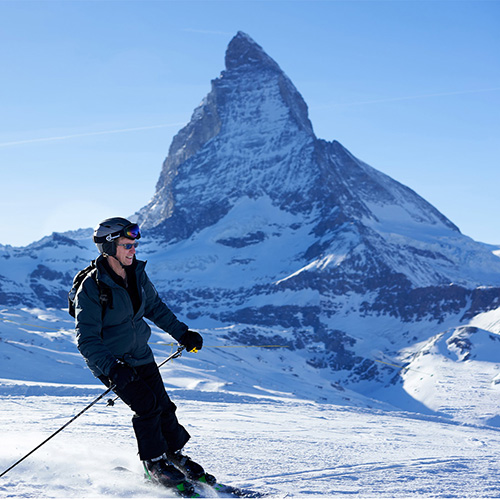 In the image, a person is skiing down a snow-covered mountain slope with a majestic, snow-capped mountain in the background.