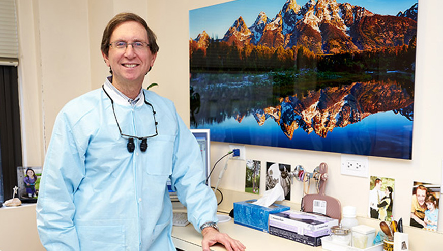 The image shows a man standing in front of a wall with a large photograph of the Grand Teton mountains. He is wearing a blue lab coat, a white face mask, and glasses, suggesting he may be a scientist or medical professional.