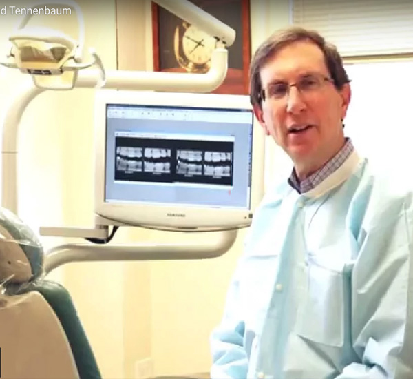 The image shows a man sitting in front of a dental chair, wearing a white lab coat and a stethoscope, with a dental monitor displaying an X-ray behind him.