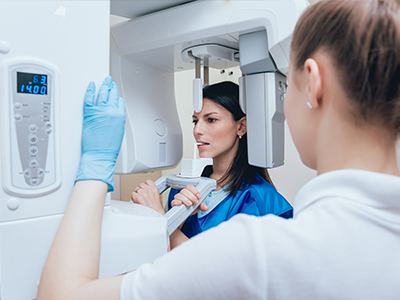 Woman in blue medical coat standing next to a large white and gray CT scanner machine, with another woman in blue gloves inspecting the device.